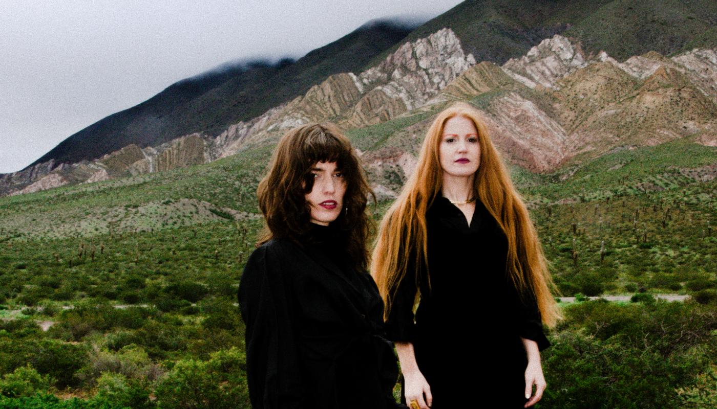 Two women, one with dark hair and one with red hair, stand in front of a green landscape. 